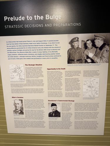 Large printed wall panel titled 'Prelude to the Bulge – Strategic Decisions and Preparations', with sub-sections 'The Strategic Situation', 'Opportunity in the South', 'Hitler's Decision', and 'Eisenhower's Controversial Strategy'. Includes maps, a photo of two senior officers, and portraits of Hitler and Eisenhower.