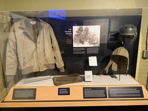 Glass display case under the heading 'THE COLDEST WINTER' (with a group photo of bundled-up soldiers). Inside: a winter combat jacket and wool sweater, folded scarf and gloves, a Christmas V-Mail card, and a US armored-forces tanker helmet plus a winter combat (tanker) cloth helmet. A row of small white labels runs along the bottom.
