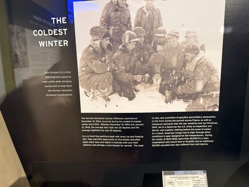 Close-up of the 'THE COLDEST WINTER' graphic inside the same case: a black-and-white photo of a cluster of bundled, snow-covered GIs huddled together making coffee, with two body-text columns below and a glimpse of a V-Mail card at the bottom edge.