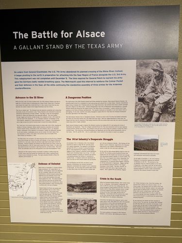 Large printed wall panel titled 'The Battle for Alsace – A Gallant Stand by the Texas Army', with sub-sections 'Advance to the Ill River', 'A Dangerous Position', 'The 141st Infantry's Desperate Struggle', 'Defense of Selestat', and 'Crisis in the South'. Two black-and-white photos at upper and lower right.