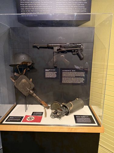 Tall vertical display case of captured German equipment. Mounted high is a German MP-40 submachine gun; below it a German Model 1942 steel helmet, an entrenching tool, a small red Nazi (swastika) flag, and a Model 1938 gas mask with its ribbed metal canister. Two black label cards are visible. A blue/black wall-text band runs across the top mentioning Christmas 1944 and 'White Christmas'.