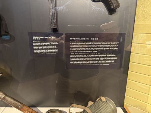 Close-up of the two main label cards in the captured-German-weapons case: 'German Model 1942 Helmet' and 'MP-40 Submachine Gun'. There is glass glare and a faint reflection of the photographer's hand, but the text is legible.
