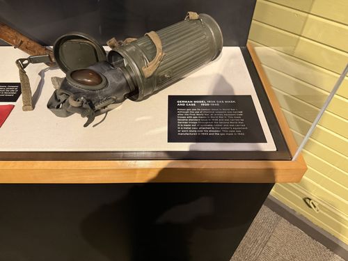 Close-up of the German Model 1938 gas mask resting in the case with its ribbed cylindrical metal carrier case, plus a black label card 'German Model 1938 Gas Mask and Case'. A leather strap and the red Nazi flag are visible at the left edge.