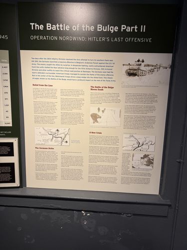 Large printed wall panel titled 'The Battle of the Bulge Part II – Operation Nordwind: Hitler's Last Offensive', mounted on a gray metal wall. Sub-sections include 'Relief from the Line', 'The Battle of the Bulge Moves South', 'The Germans Strike', and 'A New Crisis'. Maps, a tank photo, and snow-scene photos illustrate it; a partial casualty-total chart is on the left edge.