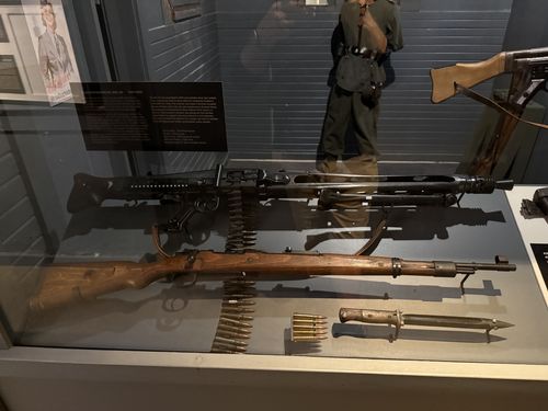 Display case of captured German weapons: a belt-fed MG 42 machine gun on its bipod with a linked ammunition belt, a Mauser bolt-action rifle below it with stripper-clipped cartridges, and a bayonet. A German uniform stands behind the glass and a folding-stock weapon hangs at upper right. The MG 42 placard is at upper left.
