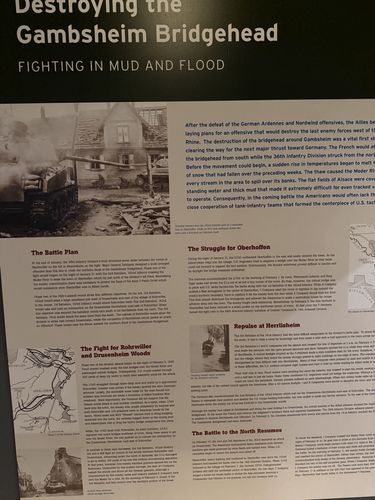 Angled photo of a large interpretive panel titled 'Destroying the Gambsheim Bridgehead - Fighting in Mud and Flood', with a destroyed-building photo, a map of the Alsace area, and multiple text sections (The Battle Plan, The Struggle for Oberhoffen, Repulse at Herrlisheim, The Fight for Rohrwiller and Drusenheim Woods, The Battle to the North Resumes).