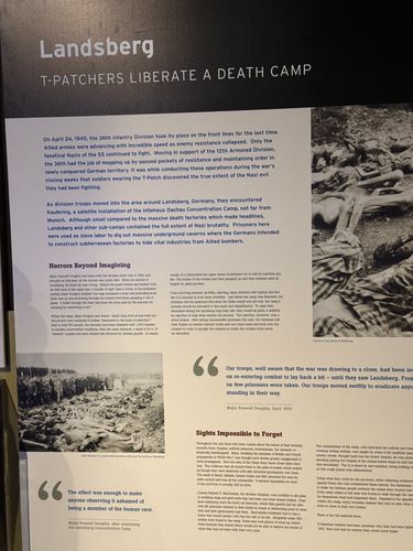 Straight-on photo of a large wall panel titled 'Landsberg - T-Patchers Liberate a Death Camp', with archival photographs of the camp and the dead, the division's discovery of Dachau satellite camps, and quotes from Major Roswell Doughty.