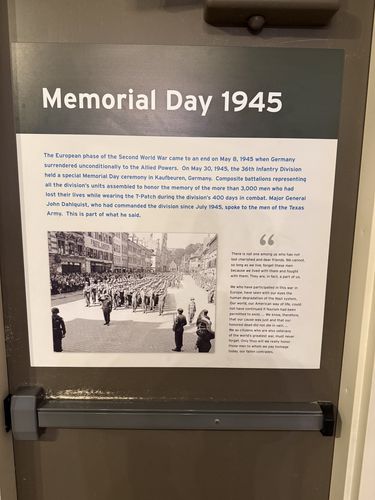 A panel mounted on a metal exit door (a push-bar is visible) titled 'Memorial Day 1945', with a photograph of the division's Memorial Day parade in Kaufbeuren, Germany, and a quote from Major General John Dahlquist.