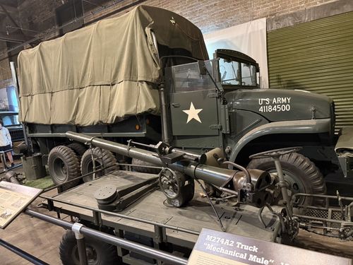 A large olive-drab canvas-topped cargo truck marked 'U S ARMY 41184500' (an M135-series 2.5-ton truck) parked against a brick wall. In the foreground sits the small M274 'Mechanical Mule' flatbed carrier with a recoilless rifle mounted on it, and a brown placard reading 'M274A2 Truck Mechanical Mule'.