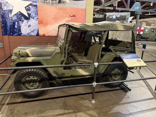 An olive-drab M151 'MUTT' military jeep with its canvas top up, displayed behind a railing. A small blue-and-tan placard beside it reads 'M151A2 MUTT 1970-2000'. A painted mural and an aircraft tail (U.S. Army) are visible in the background.