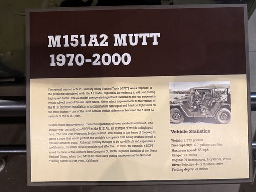 Close-up of the brown-and-tan placard titled 'M151A2 MUTT 1970-2000', with explanatory text about the jeep's roll-over problems and the Roll Over Protection System (ROPS), a black-and-white photo of the jeep, and a vehicle statistics block.