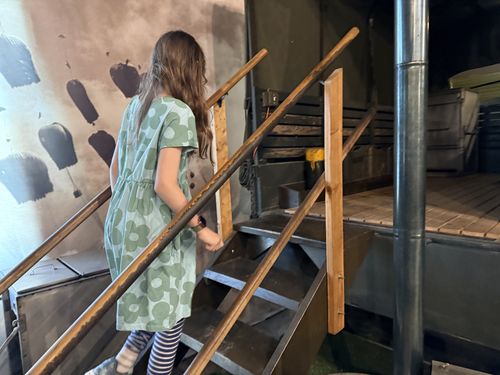 Maxine, in a green floral dress and striped leggings, climbing the wooden steps up into the open back of the M211 field-kitchen truck. The shiny immersion-heater chimney and the wooden floor of the truck bed are visible; a mural with hanging objects is on the wall behind.