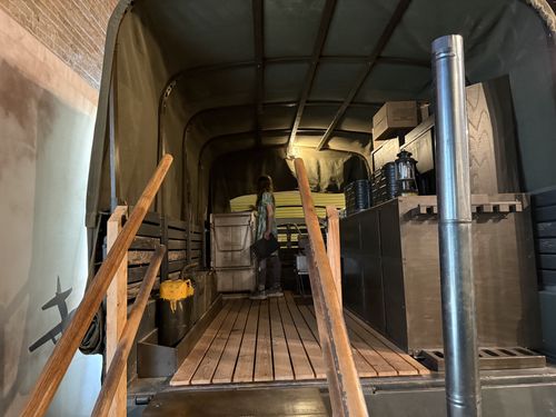 Interior view from the rear ramp of the M211 mess truck, with Maxine standing in the middle of the wooden-floored bed. Stoves, mermite food cans, a lantern, stacked supply boxes, and the tall immersion-heater chimney line the right side.