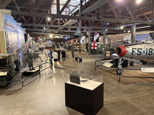 Wide view of the museum's main gallery hall: an F-84 Thunderjet jet fighter with red nose and 'FS-188' marking on the right, an olive-drab ambulance truck with a red cross, aircraft hanging from the timber-trussed ceiling, a Confederate-style flag, a uniformed mannequin on the left, and several visitors in the center.