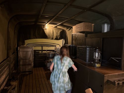 Interior of the M211 mess truck with Maxine (motion-blurred) walking toward the kitchen counter. Visible are the gasoline stoves with pots, stacked black mermite cans, a Coleman lantern, supply boxes, and the immersion-heater chimney. Maxine is blurred from movement but the kitchen detail is clear.