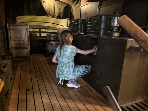 Maxine crouched on the wooden floor of the M211 mess truck, opening a cabinet door in the kitchen counter and inspecting the contents. Stacked black mermite food cans and a Coleman lantern sit on the counter above her; pots and gear are on the far side.