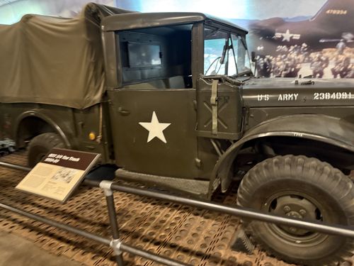 An olive-drab M37 3/4-ton Dodge truck with canvas cargo cover and a white star on the door, marked 'U S ARMY 2384091'. A small angled placard ('M37 3/4 Ton Truck 1950-80') sits on the floor in front of the cab; a wartime mural is behind.