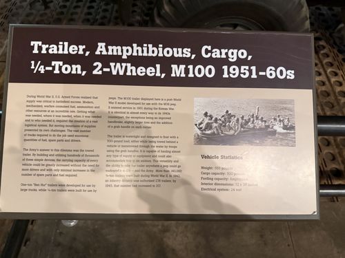 A brown-and-tan placard titled 'Trailer, Amphibious, Cargo, 1/4-Ton, 2-Wheel, M100 1951-60s', with text about Army logistics and the floating M100 trailer, a photo of soldiers crossing water in the trailer used as a boat, and a vehicle statistics block.