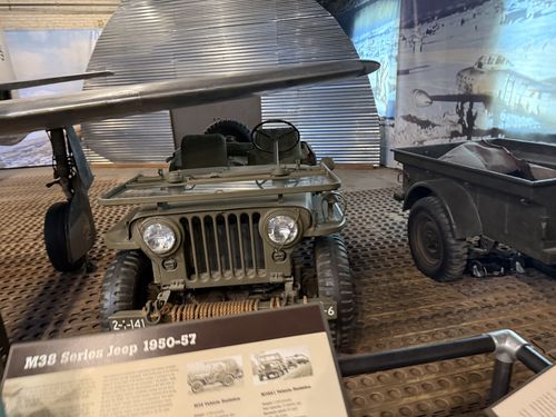 The M38 jeep itself, photographed head-on. Olive-drab open jeep with a folded windshield, a coil of rope on the front bumper, and a small trailer hitched behind it. A light-plane wing crosses overhead and Korean-war photo banners line the wall behind. The M38 placard is visible at the bottom edge.