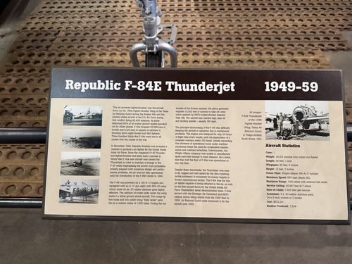 Straight-on view of the brown 'Republic F-84E Thunderjet 1949-59' placard with the landing gear of the actual jet visible above it. Includes black-and-white photos and a full Aircraft Statistics block.