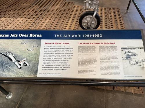 Angled shot of the left end of the blue 'Texas Jets Over Korea — THE AIR WAR: 1951-1952' banner placard, with the F-84's nose wheel above it. Sections 'Korea: A War of Firsts' and 'The Texas Air Guard Is Mobilized' plus photos of jets and a pilot.