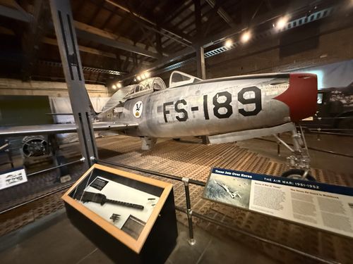 Wide hero shot of the silver Republic F-84E Thunderjet (tail markings 'FS-189') with its red nose ring, parked in the hangar gallery under wooden roof trusses. A glass display case holding a machine gun sits in the foreground, with the Korea air-war banner placards on the right rail.