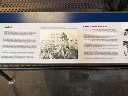 Angled shot of the right-center of a blue Korean War banner placard, showing the 'Inchon' and 'China Enters the War' sections with a black-and-white photo of Marines climbing a seawall ladder at Inchon.