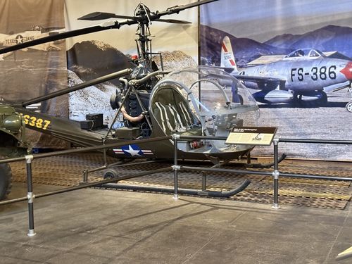 An olive-drab OH-23 'Raven' light observation helicopter with an open bubble canopy and a tall exposed rotor mast, displayed on skids inside a fenced area. Behind it, a wall mural shows a Thunderjet marked 'FS-386'; a small placard sits on the cockpit rail. A tank fender marked '...6387' is at far left.