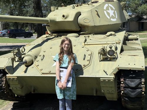 Outdoor lawn shot: Maxine in a teal floral dress and glasses stands smiling in front of a large olive-drab light tank with a white star on the turret, holding her scavenger-hunt papers. Cars and a building are visible behind in the parking area.
