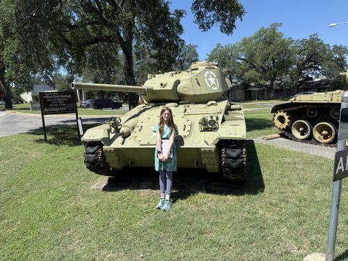 Wider outdoor shot of the same scene: Maxine standing in front of the olive-drab light tank on the museum's front lawn under blue sky, with a second armored vehicle, an information sign, and trees in the background.
