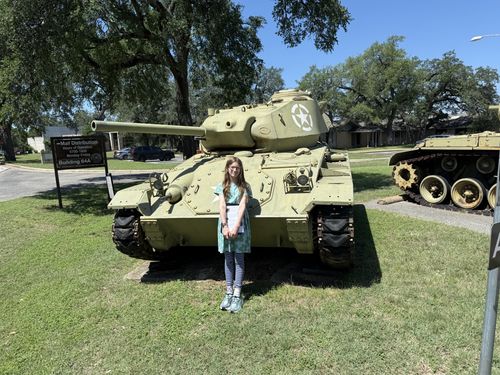 Maxine stands in front of a green American light tank in the outdoor vehicle park, the long gun barrel angled left overhead and a white US star on the turret. A post directional sign and a second tank are visible at right; oak trees and blue sky behind.