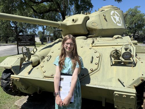 Closer landscape-format portrait: Maxine in a teal dress in front of the light tank, the 'Mail Distribution / Hours of Operation 0700-1600 Monday-Friday / Building 64A' post sign clearly readable at left, white star on the turret at right. Crisp and detailed.