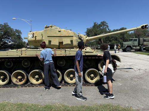 Candid shot of a separate, weathered sand/desert-painted tank (yellow tan, overlapping road wheels suggesting an M47/M48-type medium tank) under a streetlight; three teenage visitors lean against and peer over the hull. In the background are oak trees, an M35-type military cargo truck, and another visitor.