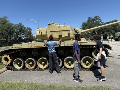 Wider candid of the same sand-painted tank a moment later, showing the full row of overlapping road wheels, the long gun barrel, and the three teens alongside; an M35 cargo truck and more visitors are in the background. Good detail of the running gear.
