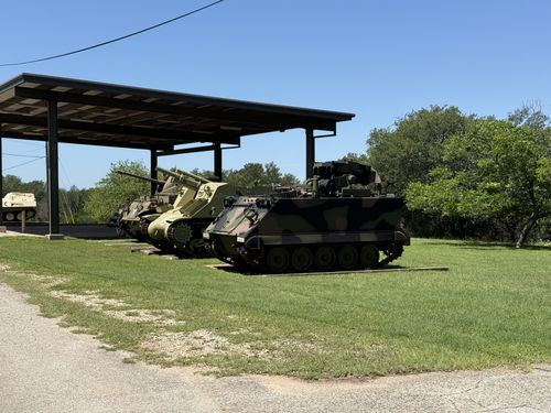 A covered open-sided display shelter on the museum grounds holding several tracked vehicles in a row: an open-topped self-propelled gun, a smaller tracked vehicle, and an M113-family armored personnel carrier in green/brown/black MERDC woodland camouflage parked on the grass out front. Bright sky, brush and trees behind.
