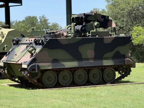 Close side view of the camouflaged M113-family APC in MERDC woodland pattern, showing its five road wheels, track, trim vane on the front, and a tall mast/launcher fitting on top. To the left, another tracked vehicle under the shelter is marked 'The Texas Special.'